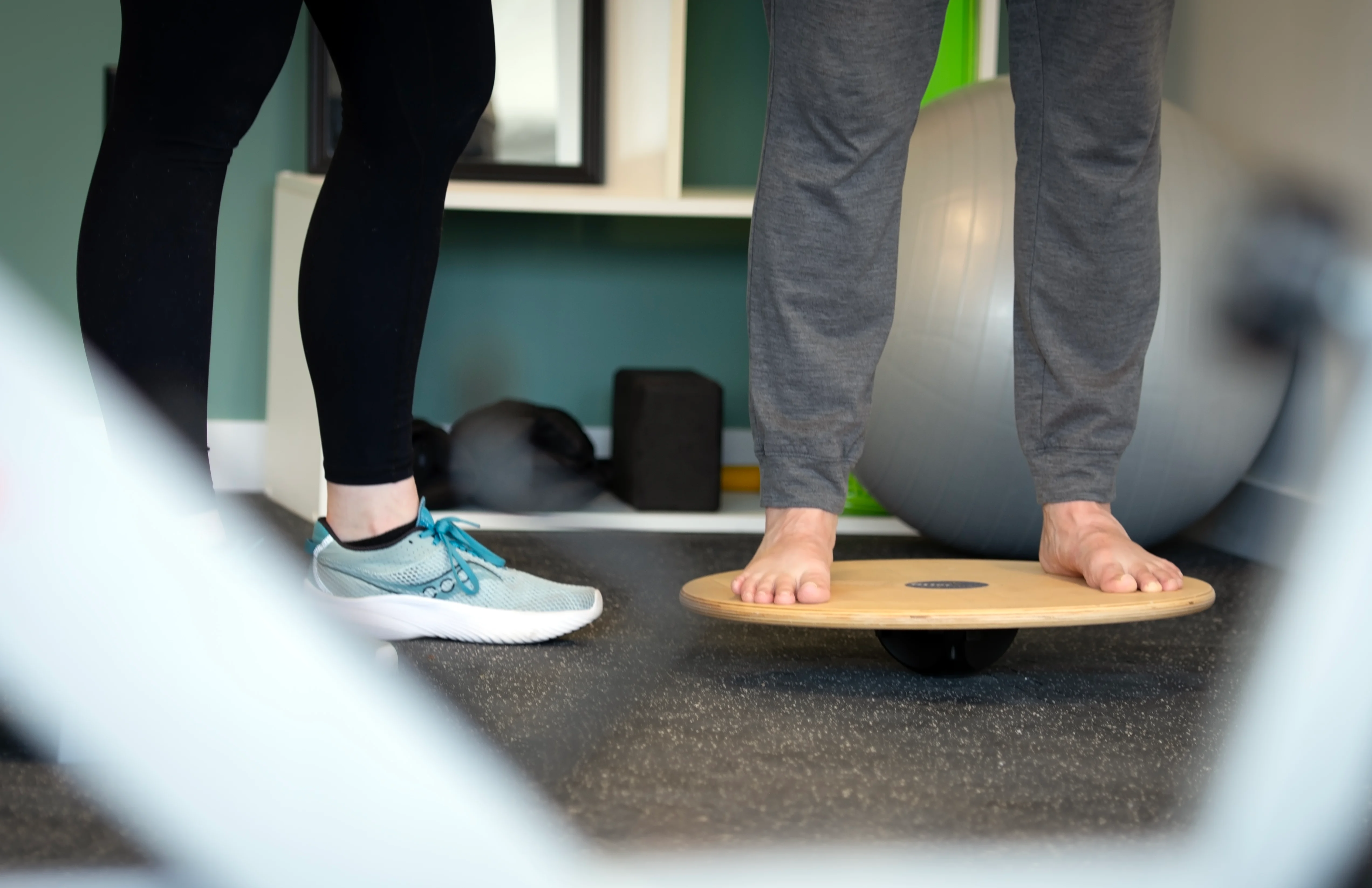 A patient balancing barefoot on a wooden wobble board beside an exercise ball, with a therapist standing close by.