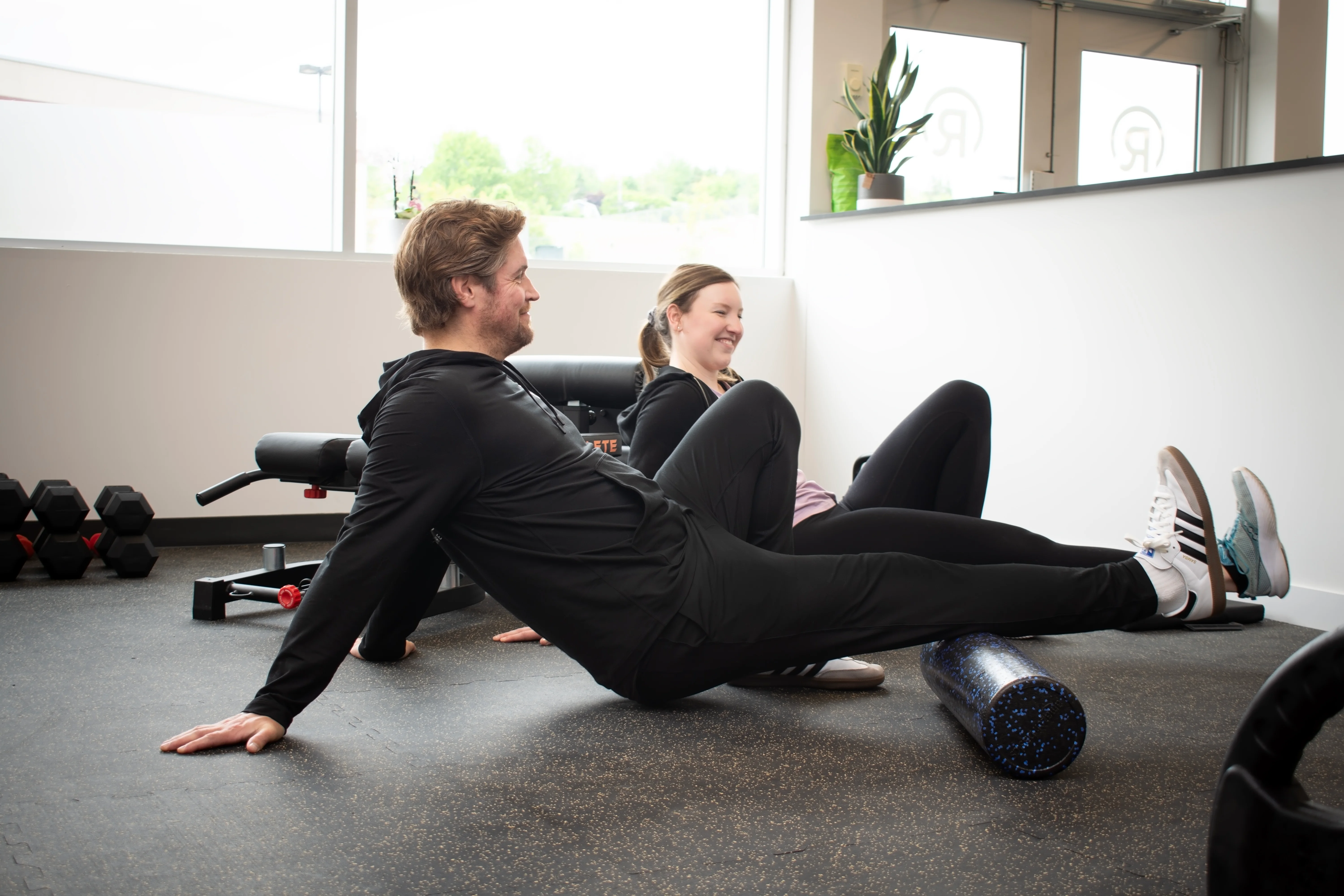 Two people sitting on the gym floor at Radius beside the front windows, with dumbbells and a foam roller nearby.