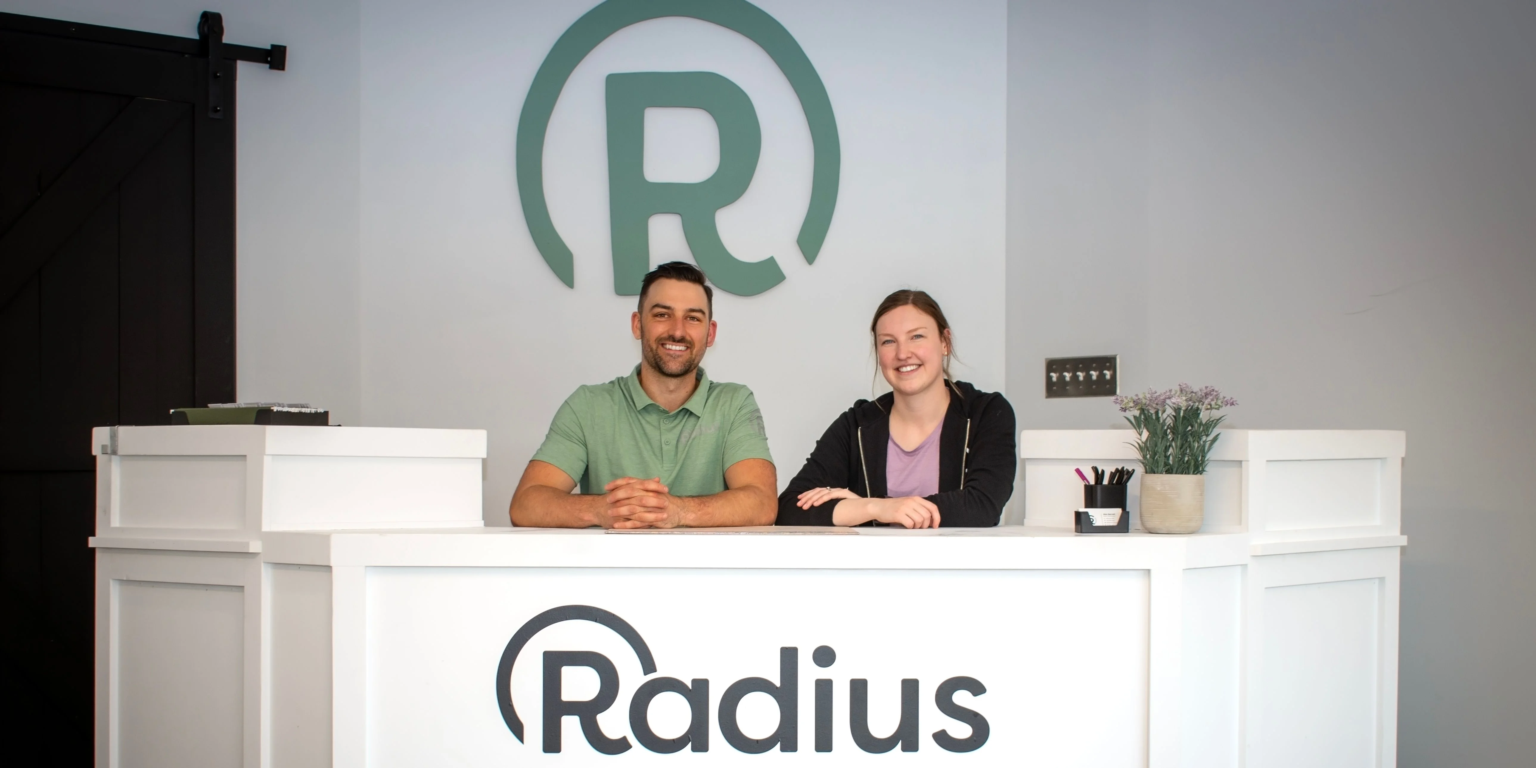 Two Radius team members behind the clinic's white reception desk, with the Radius logo on the desk front and on the wall behind them.