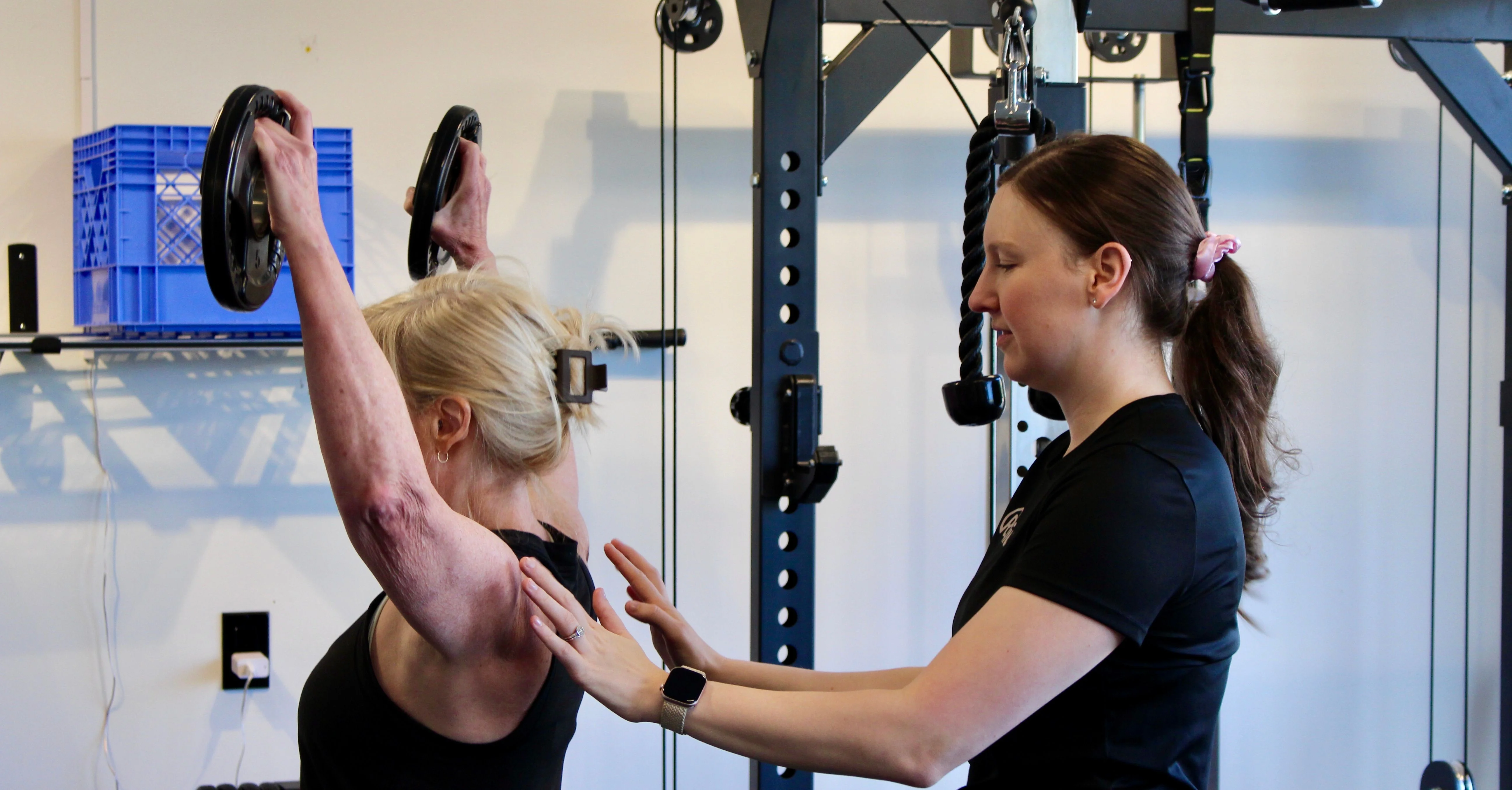 A Radius physiotherapist steadying a patient's shoulder blade as she presses two weight plates overhead.