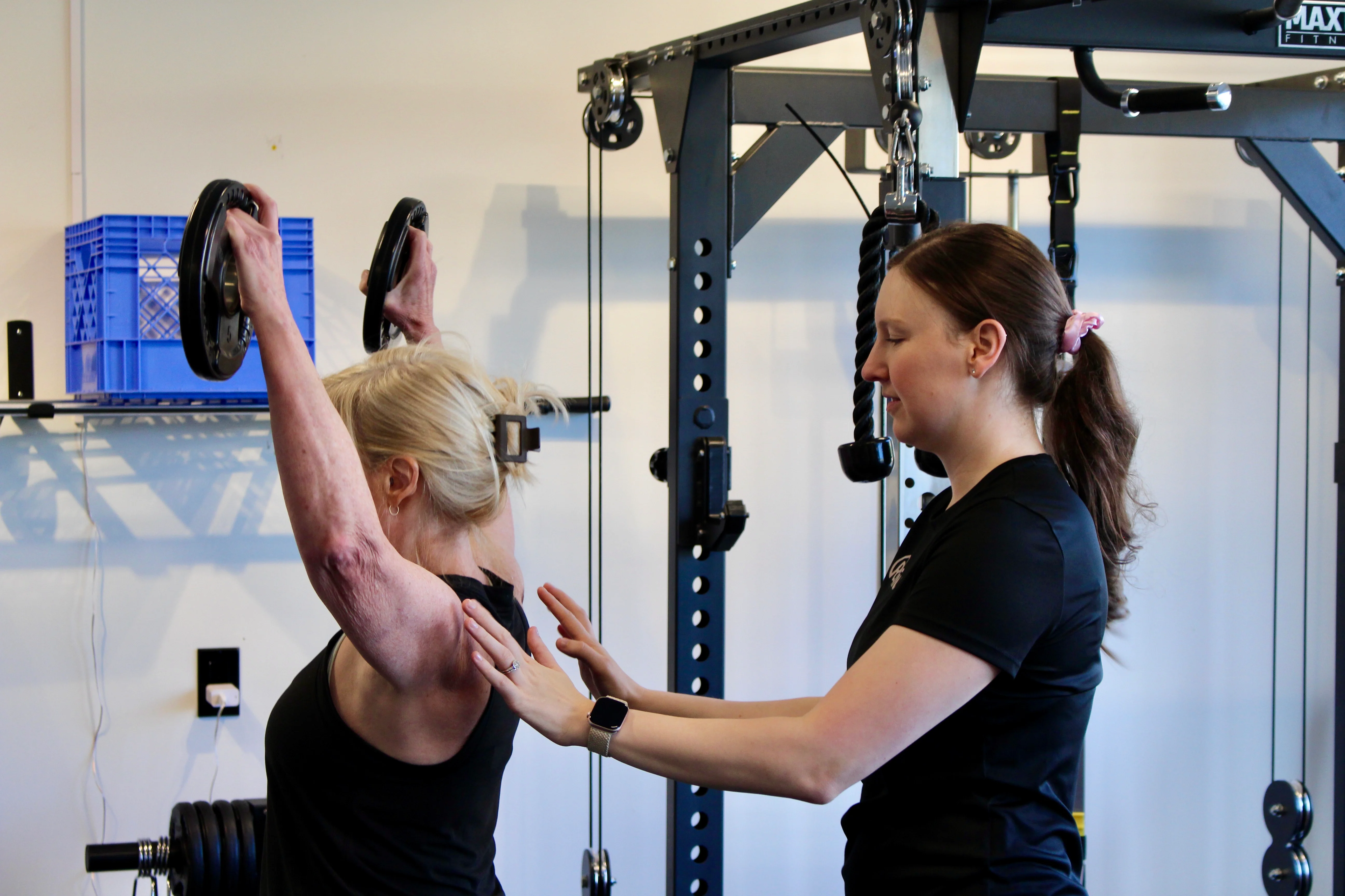 A Radius physiotherapist steadying a patient's shoulder blade as she presses two weight plates overhead.