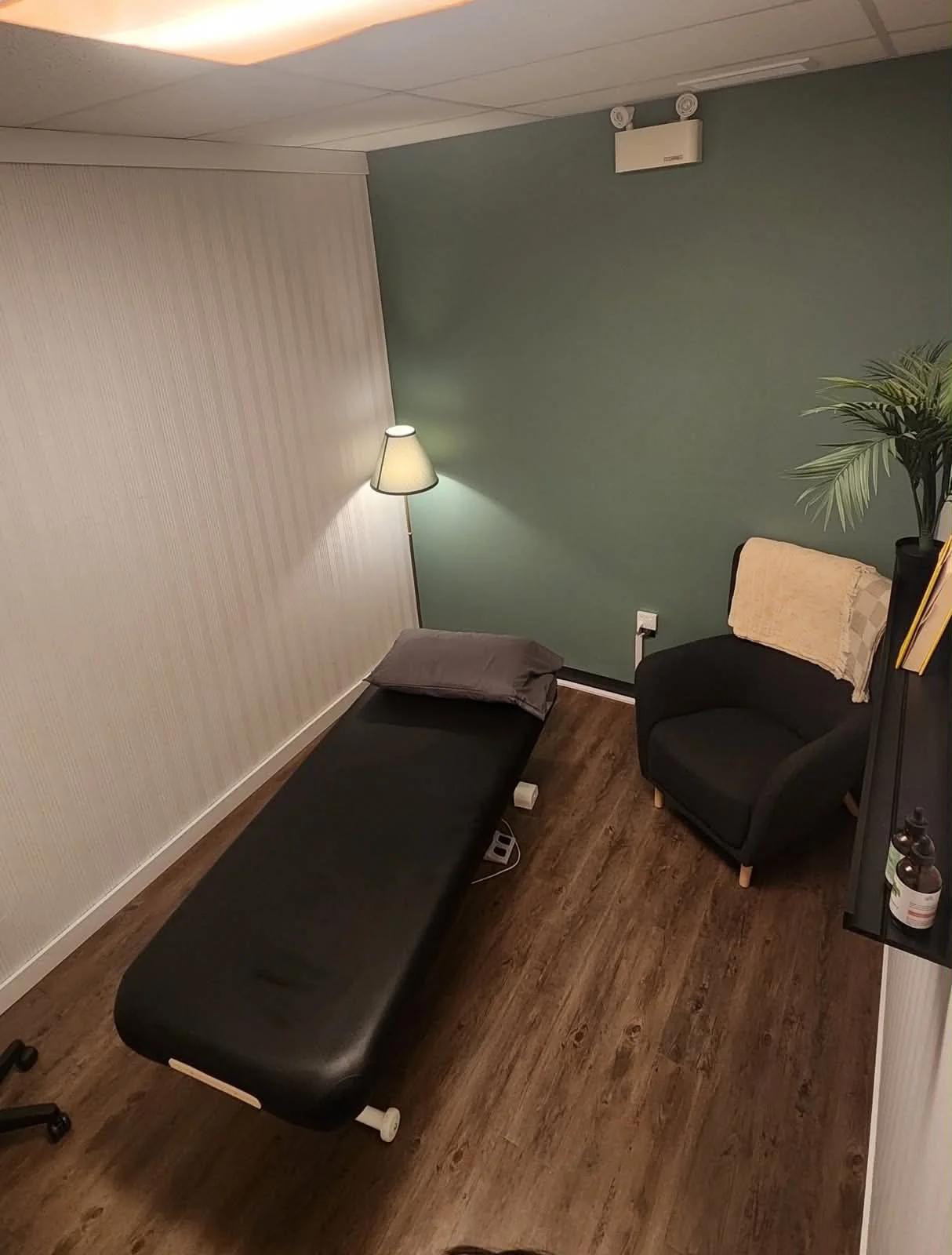 A quiet treatment room with a padded table, an armchair and a lamp against a sage green wall.